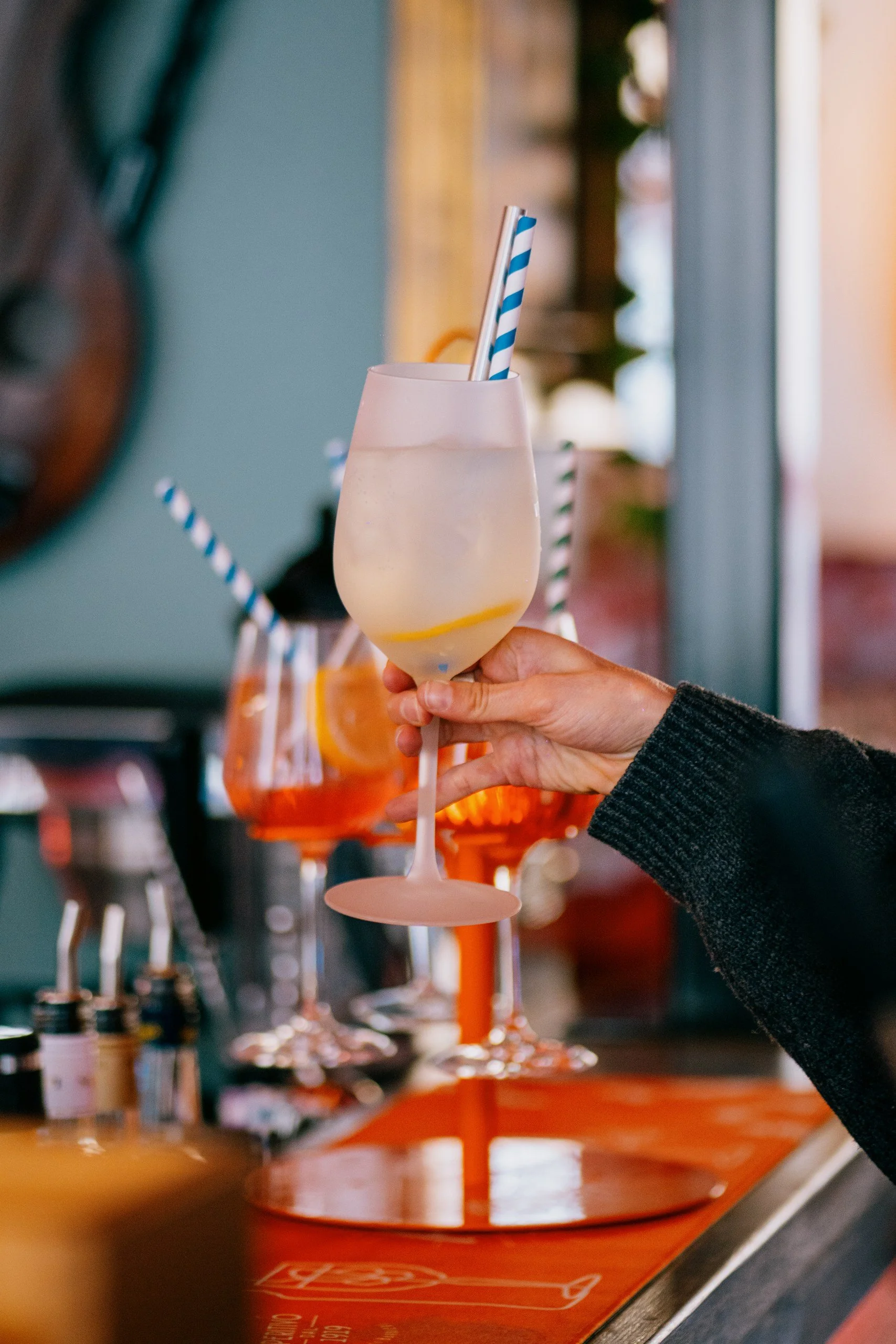 A hand holding a large, pale yellow cocktail with ice and lemon slices in a tall glass with blue-striped straws, with other similar drinks and bar supplies in the background.