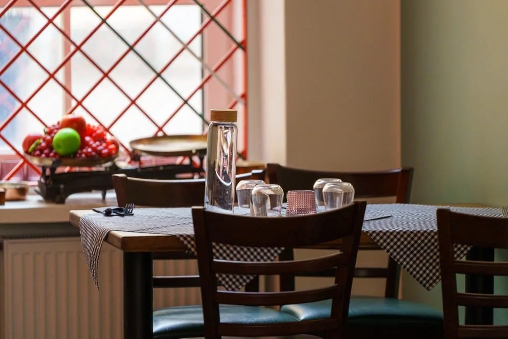 A dining table set with glasses, a water pitcher, and a checkered tablecloth in a restaurant with a window ledge holding a fruit display.