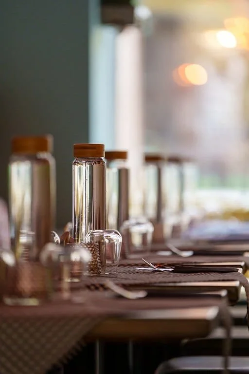A row of glass jars with wooden lids on a dining table set for a meal, with utensils and napkins. The background shows a window with sunlight.